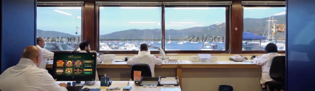 Una vista panoramica all'interno del laboratorio di orologeria Locman a Portoferraio. Un mastro orologiaio in camice bianco è seduto a un banco da lavoro professionale, concentrato su un movimento meccanico. Dalla grande finestra si gode una spettacolare vista sul porto affollato di barche a vela e sulla costa collinare, unendo la tradizione orologiera alla bellezza dell'Isola d'Elba.